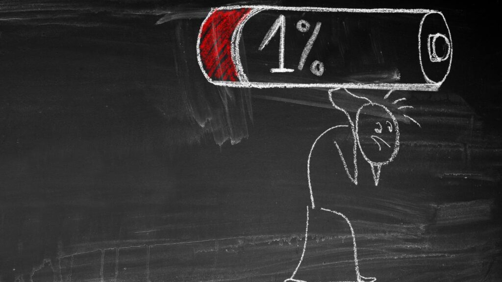 Rysunek kredą na tablicy przedstawiający wyczerpaną postać dźwigającą baterię z poziomem naładowania wynoszącym 1%, symbolizujący brak energii lub wypalenie.
This description captures the metaphorical and visual meaning of the image