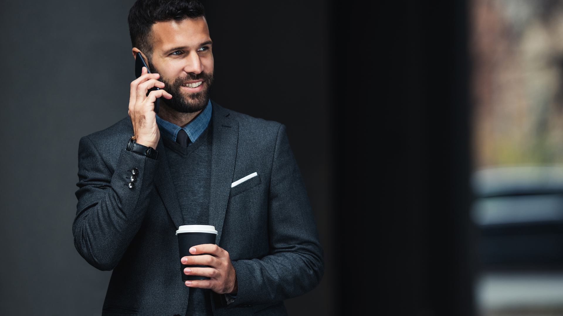 Uśmiechnięty mężczyzna w garniturze rozmawiający przez telefon z potencjalnym klientem, trzymający kubek z kawą, stojący w nowoczesnym, miejskim otoczeniu.