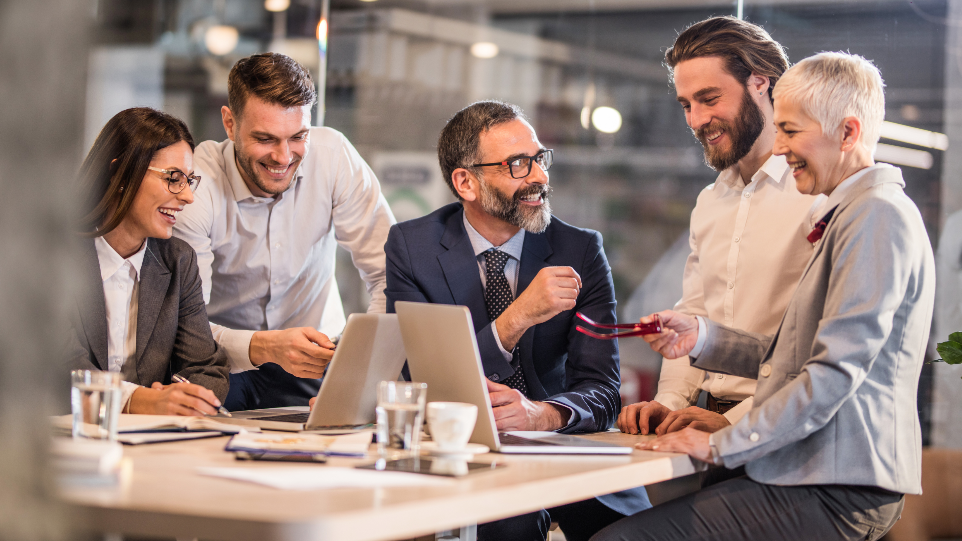 Pięcioro profesjonalistów w formalnych strojach siedzi przy stole w nowoczesnym biurze, zaangażowanych w żywą i współpracującą dyskusję. Wszyscy się uśmiechają i wyglądają na skupionych na wspólnym projekcie, pracując przy laptopie. Atmosfera jest pełna pracy zespołowej i pozytywnej energii, w grupie znajdują się zarówno mężczyźni, jak i kobiety, a także osoby młodsze i starsze