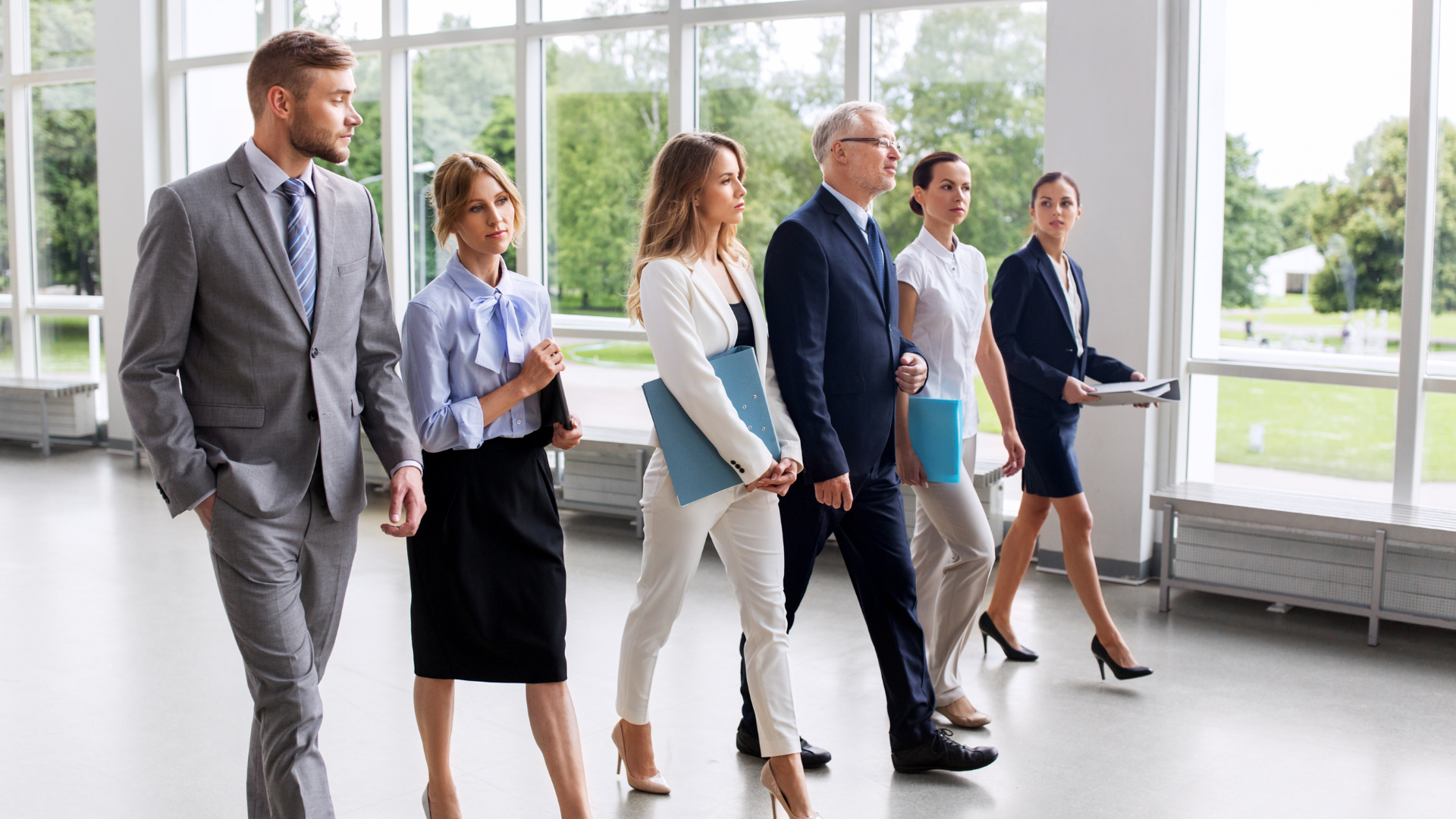 Grupa profesjonalistów w formalnych strojach, idących razem przez nowoczesne biuro z dużymi oknami, trzymając dokumenty i teczki.